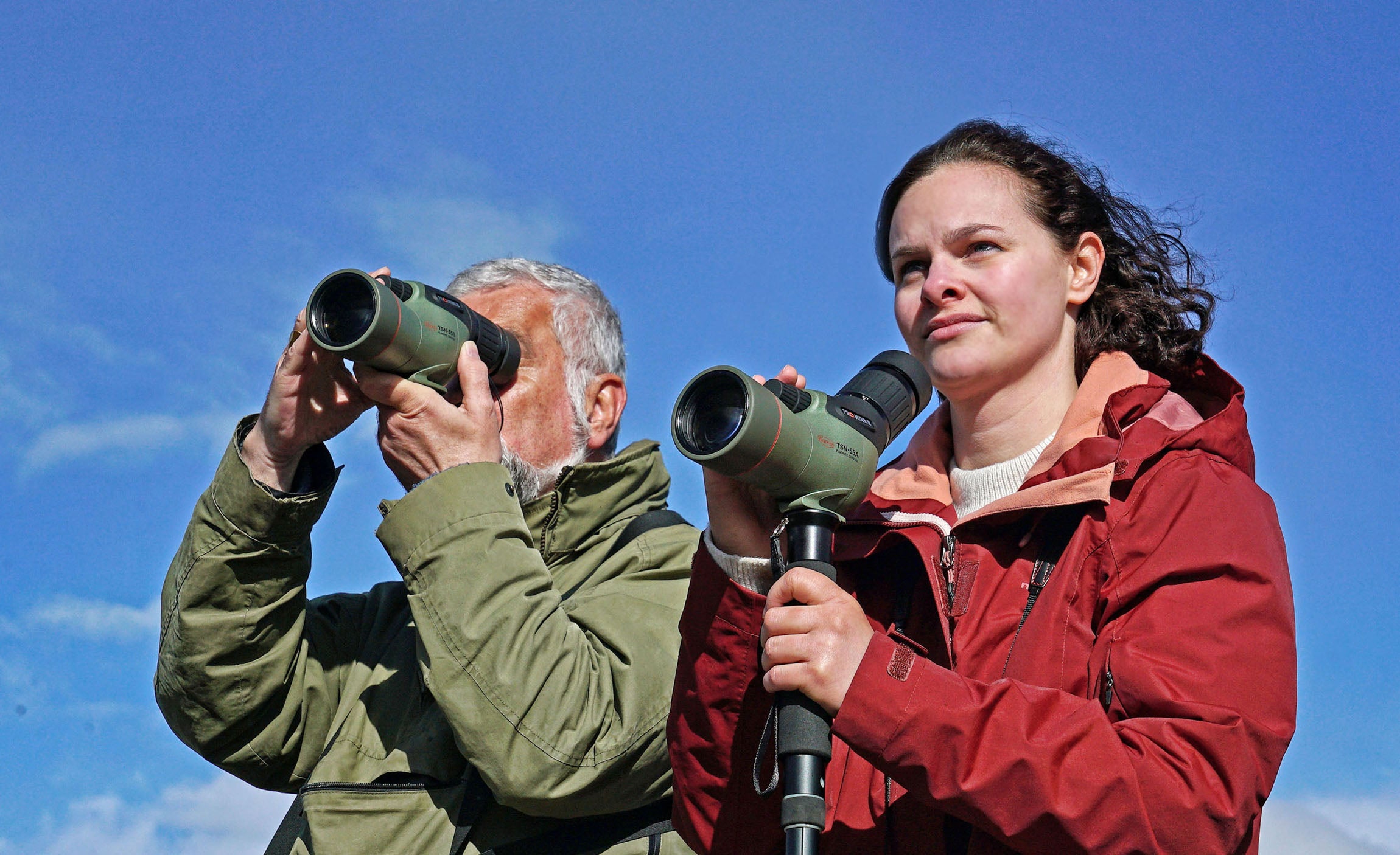 Two enthusiasts using the Kowa TSN-55S PROMINAR Straight Spotting Scope against a blue sky, enjoying outdoor observations.